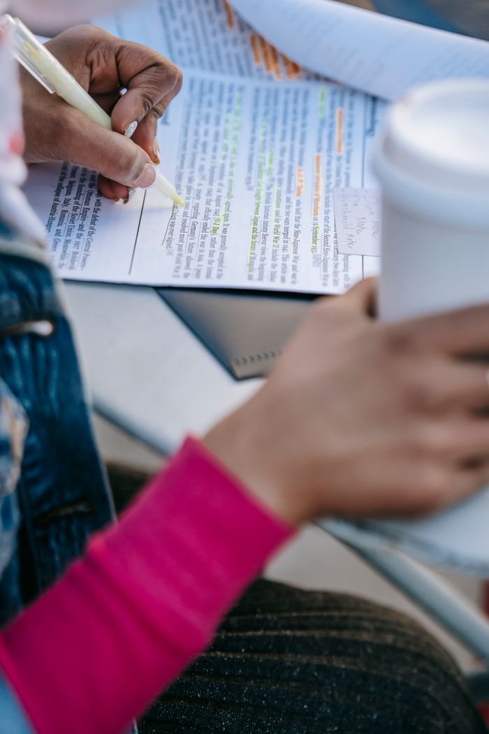 From above of crop anonymous student sitting at table with takeaway drink and using marker for highlighting important information in document in daytime