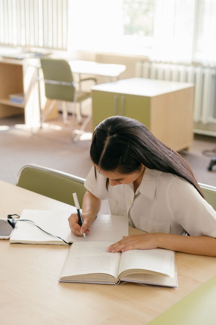 services-01 Young woman writing in a notebook, studying at a library table.